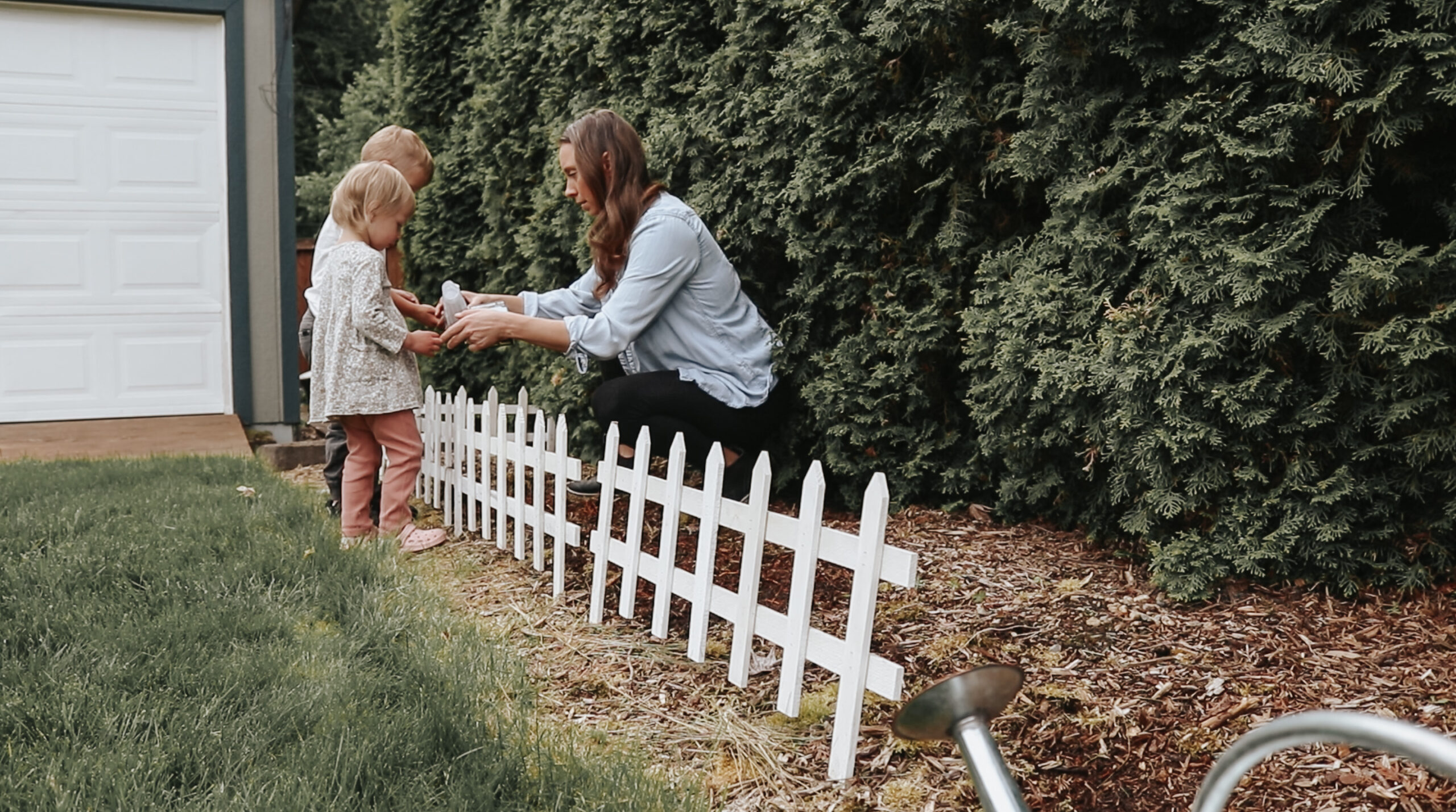 Christian homemaker shows her children something from the garden with a white picket fence in front of the garden.